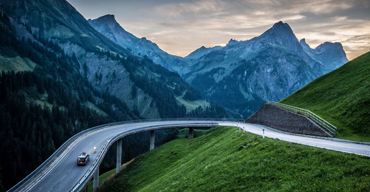 Titelbild des Race Around Austria: Kurve am Hochtannbergpass mit Radfahrer und Pace Car auf einer Brücke
